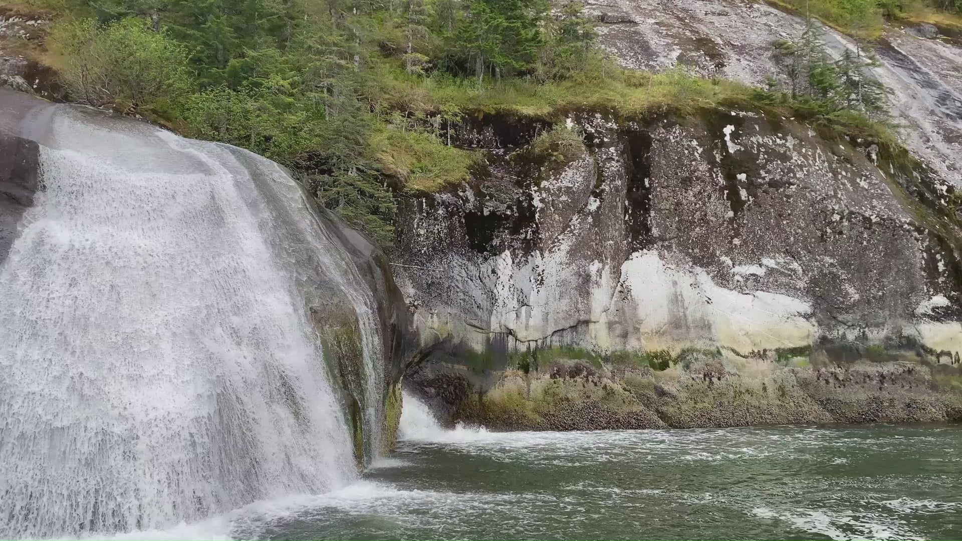 Scenic waterfall cascading over smooth rock into a forest-lined river, surrounded by mossy cliffs and evergreen trees.